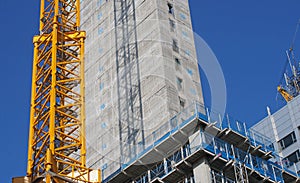 Close up of a large urban construction site with a yellow tower crane casting a shadow on a large concrete building and safety