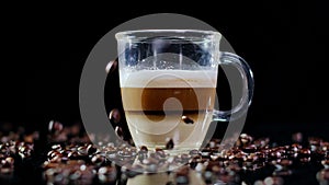 Close-up latte in glass cup with roasted coffee beans falling in slow motion on table. Closeup tasty freshly-brewed
