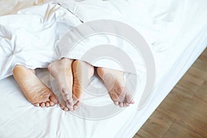 Close up legs feet of two Lovers couple sleeping side by side embracing under blanket white sheets in bed at home on holiday conc