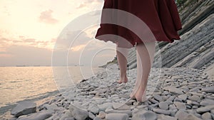 Close-up of the legs of a young girl with a red dress from behind walking on a rocky beach the sea coast at sunset. A