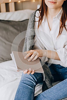 Young girl dressed in beige top sitting on bed with legs crossed, holding open notebook on knees, writing her ideas, future plans