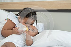 Close up of little adorable African older sister feeding newborn baby milk bottle and kissing with love and care while infant