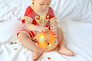 Close up little Asian baby boy in traditional Chinese dress putting some coins into a piggy bank sitting on bed at home. Kid
