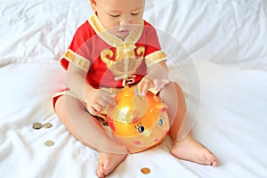 Close up little Asian baby boy in traditional Chinese dress putting some coins into a piggy bank sitting on bed at home. Kid