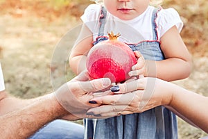 Close up little baby girl and her parents holding pomegranate fruit in sunset garden. Happy Family and fertility concept. Selectiv