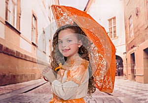 Close up of little beautiful curly girl wearing a beautiful colonial costume and holding an orange umbrella in a blurred