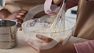 Close up of little black girl cooking pastry with mother, woman adding flour and daughter whisking dough at kitchen