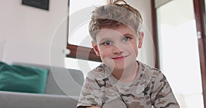 Close up of a little boy drawing on the floor, who looks to the camera and smiles shyly Grey sofa and balcony door in