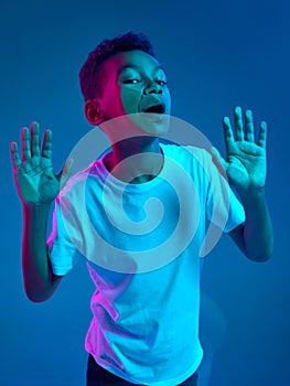 Close-up of little boy leaning against transparent glass by cheek isolated on blue background in neon light. Funny face