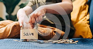 Close up of Little child kid boy hands grabbing and putting stack coins in to glass jar with save label. Donation