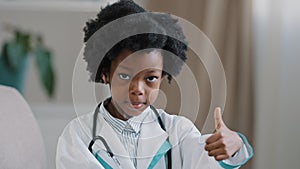 Close-up little cute kid girl in medical gown looking at camera posing indoors smiling pretending be doctor plays nurse