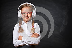 Close-up of a little girl cute student in headphones and glasses on the background of a school black board.