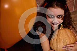 Close-up little girl with face art makeup, looks like a witch, enchantress hugging pumpkin, smiles at camera. Halloween