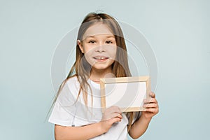 Close up little girl holding white blank frame posing isolated on pastel blue background in studio. Copy space mockup