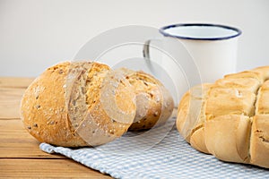 Close-up of loaf of rustic bread and rolls on cloth and wooden table with knife and cup, white background,