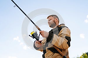 Close-up low-angle view of bearded fisherman holding casting rod wearing raincoat standing on bank waiting for bites on