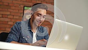 Close-up low-angle view of cheerful young man in headphones typing on laptop keyboard and talking using video call