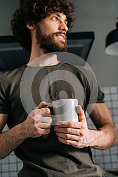 Close-up low-angle view of pensive young man holding in hand cup with morning coffee, thoughtful looking away, standing