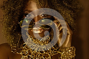 Close-up of magic golden african american female model in massive sunglasses with bright glitter makeup, glossy