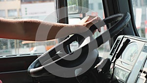 Close up male arm of lorry driver holds a big steering wheel while riding a truck at sunny day. Trucker driving to