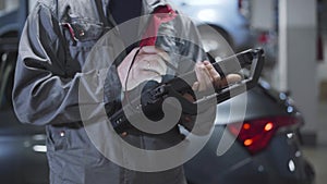 Close-up of male Caucasian hands using computer in car repair shop. Professional male auto mechanic examining automobile