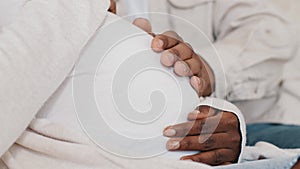 Close-up male and female hands stroking pregnant belly of woman in white clothes, African American unrecognizable couple