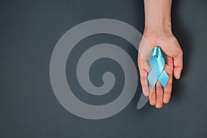 Close up of male hand holding blue ribbon awareness on grey background wall in studio with copy space for promotion content. Men`