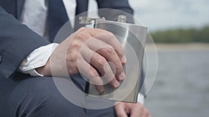 Close-up of male hand holding flask with alcohol outdoors. Unrecognizable Caucasian businessman in formal suit drinking