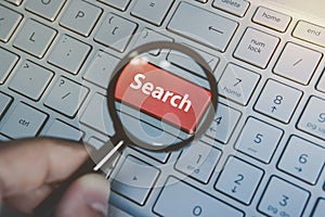 Close up of male hand holding magnifying glass above the laptop red key Search. Concept of internet searching. Enlarging