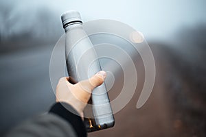 Close-up of male hand holding reusable steel thermo water bottle on blurred perspective outdoors background.