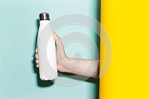 Close-up of male hand holding steel reusable thermo water bottle of white with mockup, between two backgrounds.