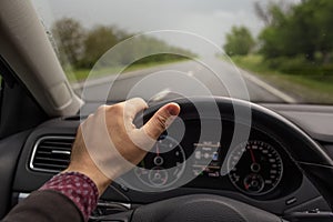 Close-up of male hand holding steering wheel, driving the car. Background of blurred road in rainy day. Travel concept.