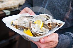 Close up male hand holding take away food tray with fresh opened oyster and lemon slices at street food market, festival, event. S