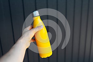 Close-up of male hand holding thermo bottle of yellow color on background of black metal fence