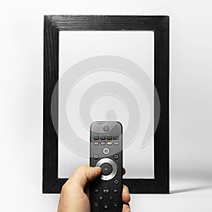 Close-up of male hand holding TV remote against black empty frame on the background of white. Artwork collage concept.