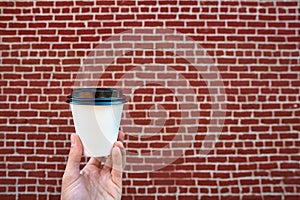 Close up of male hand holding white paper cup of coffee, against a red brick wall. Enjoying coffee to go. Fast food