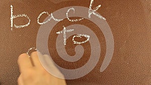 Close-up of a male hand writes the words back to school in chalk on a brown blackboard.