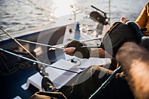 Close-up of male hands in gloves pulling rope of sailboat while mooring yacht and sitting with friends on deck.