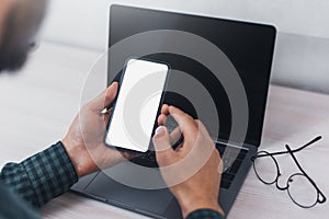 Close-up of male hands, holding smartphone on background of laptop on desk, with mockup.