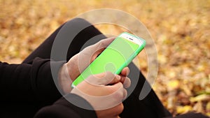 Close up male hands are holding smartphone with green screen on a background of autumn leaves