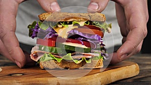 Close-up of male hands taking a tall sandwich with ham, cheese and vegetables from a wooden tray