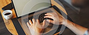 Close-up of male hands typing on laptop on wooden table near coffee cup and smartphone; panoramic banner, top view.