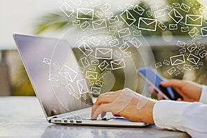 Close up of male hands using smartphone and laptop at desktop with creative drawn email icons on blurry outdoor background.