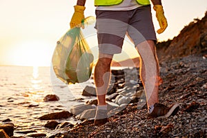 Close up of male legs of volunteer walking on wild coast holding plastic bag full of garbage. Concept of ecological