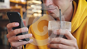Close-up of male in yellow hoodie using smartphone touchscreen in modern cafe. And drinking milkshake or smoothie