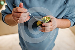 Close up of man eating avocado while standing in modern home kitchen. Slimming and healthy lifestyle concept