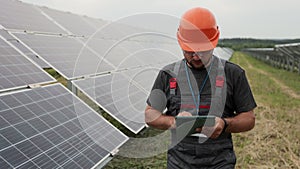 Close up of man engineer in working clothes walks on renewable energy station holding digital tablet and checking solar