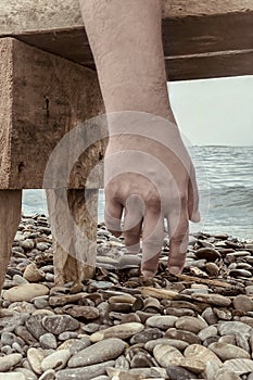 Close up man hand hanging from a beach bench, pebbles and sea on background. Atmosphere of total exhausting, prostrate, solitude,