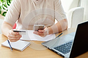 Close up of man hand holding credit card and paying purchase with smart phone at office, copy space. Technology, banking, business