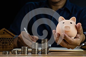 Man hand holding pink piggy bank with stacks of coins and writing for account save money. Planning step up, saving money for